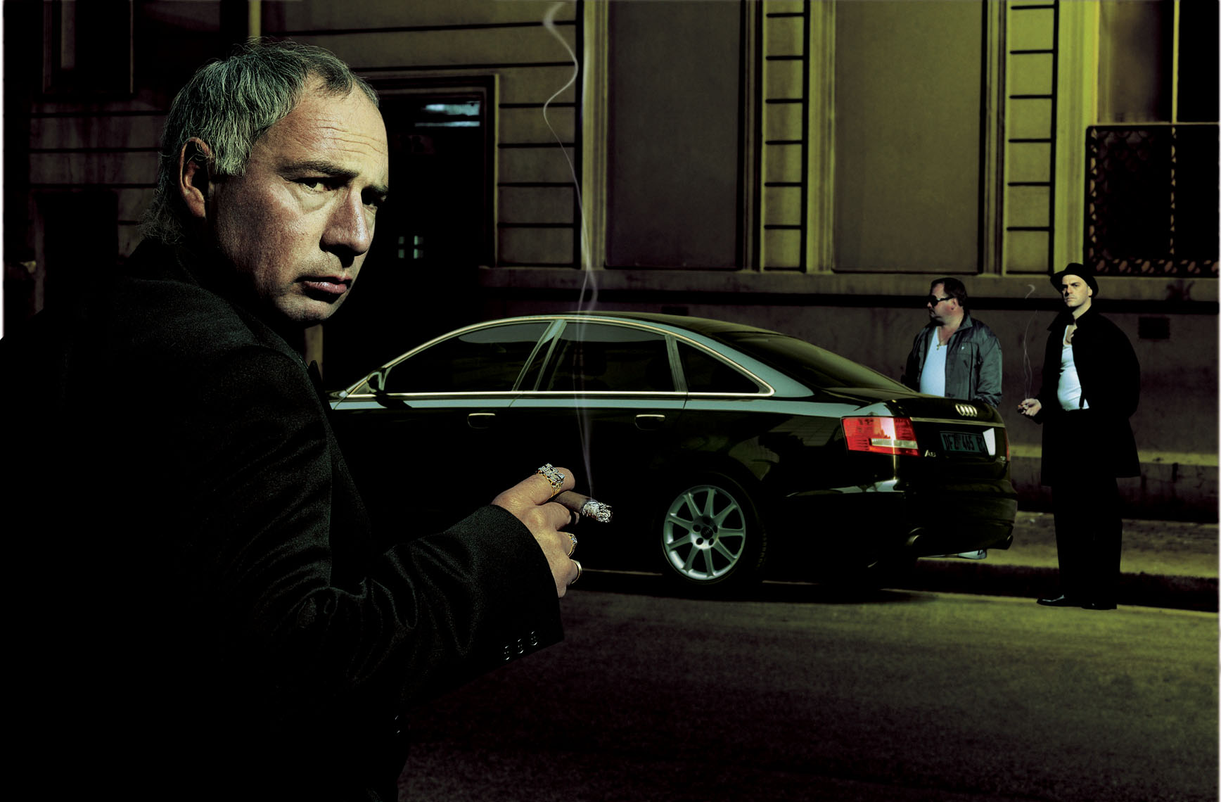 A mafia boss smoking his cigar across the road from where a black Audi A6 is parked. 
