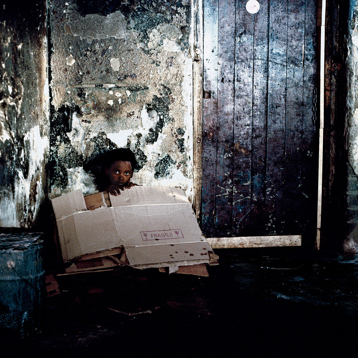 A  portrait of a street kid sitting next to a door, covered by boxes to keep warm,  in a downtown location. 
