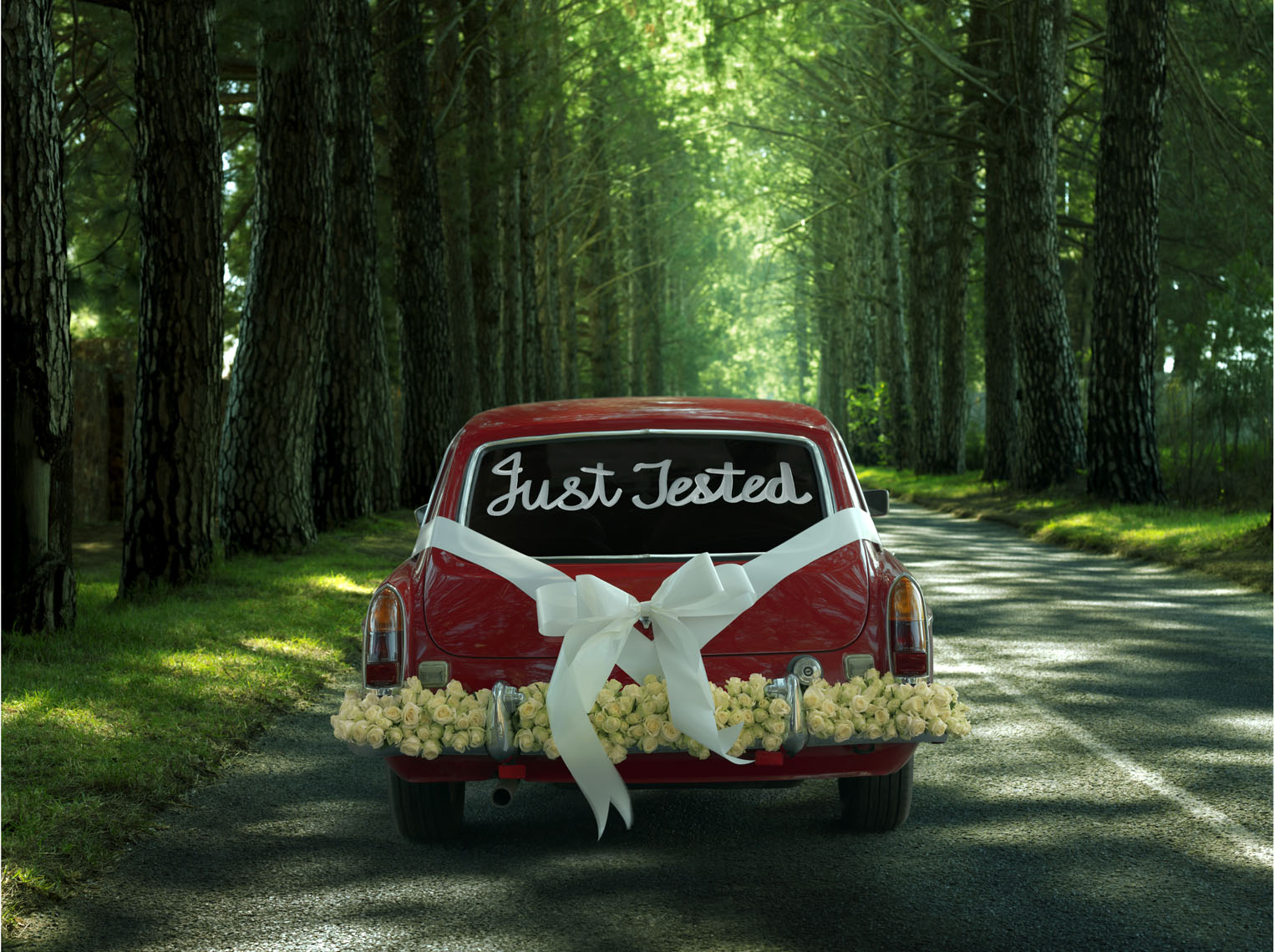 A red vintage car, with a sign reading "Just Married", decorated with ribbon and flowers driving down a country lane. 