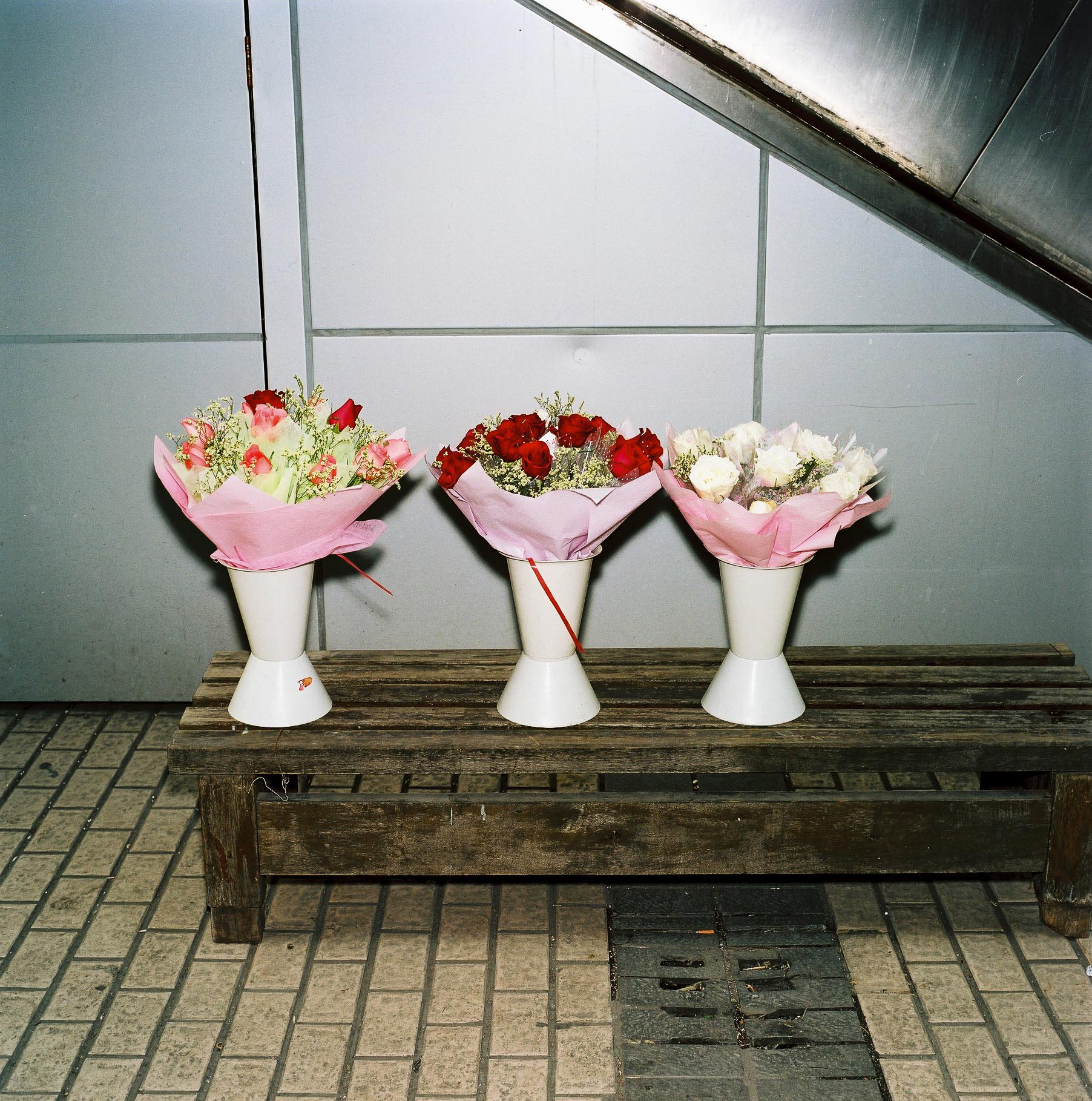 Three flower bouquets  wrapped in cellophane lined up on a wooden bench in front of an elevator. 