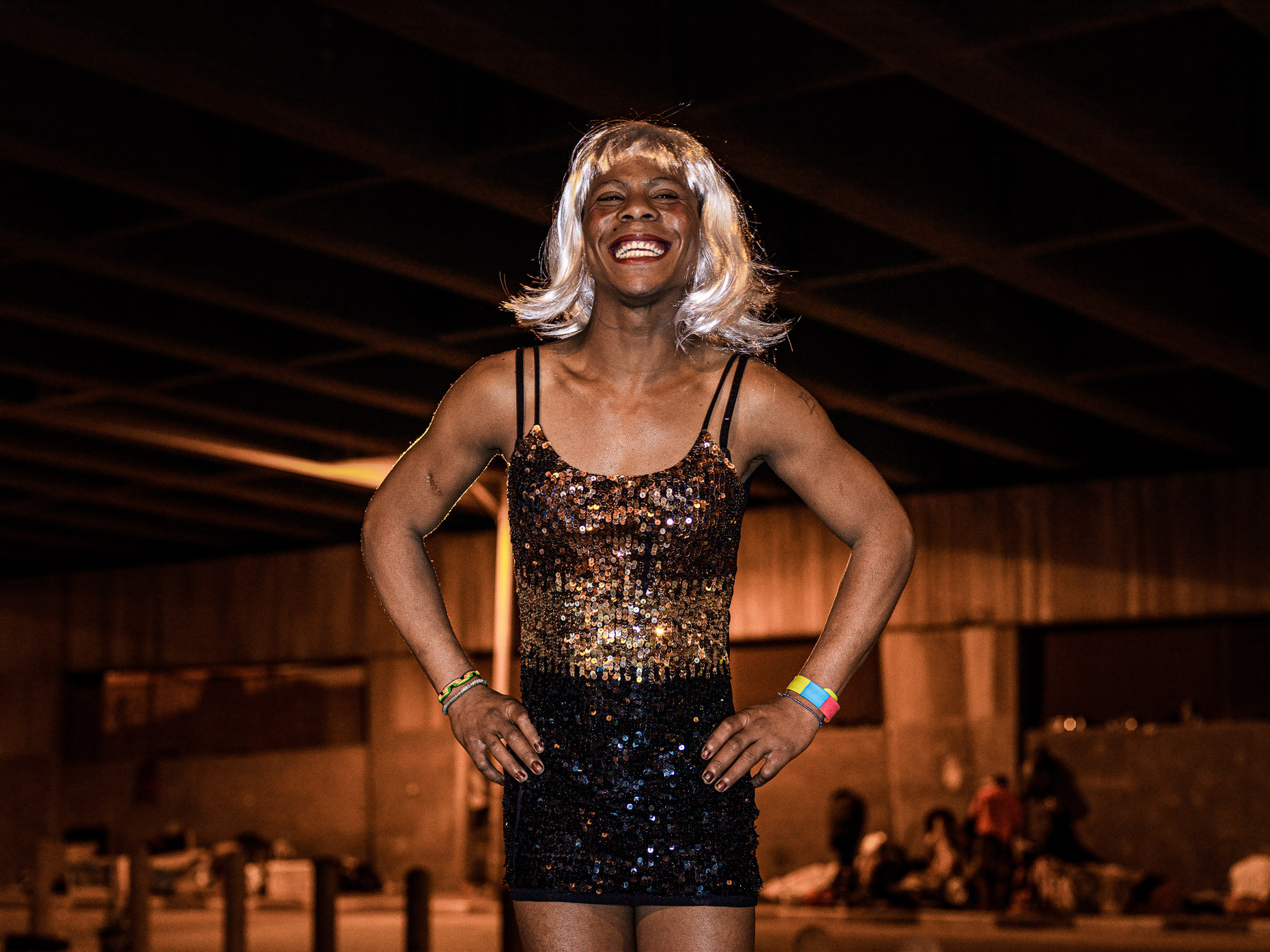  A trans woman with blonde wig laughing and posing for her portrait, under the bridge she calls home. 