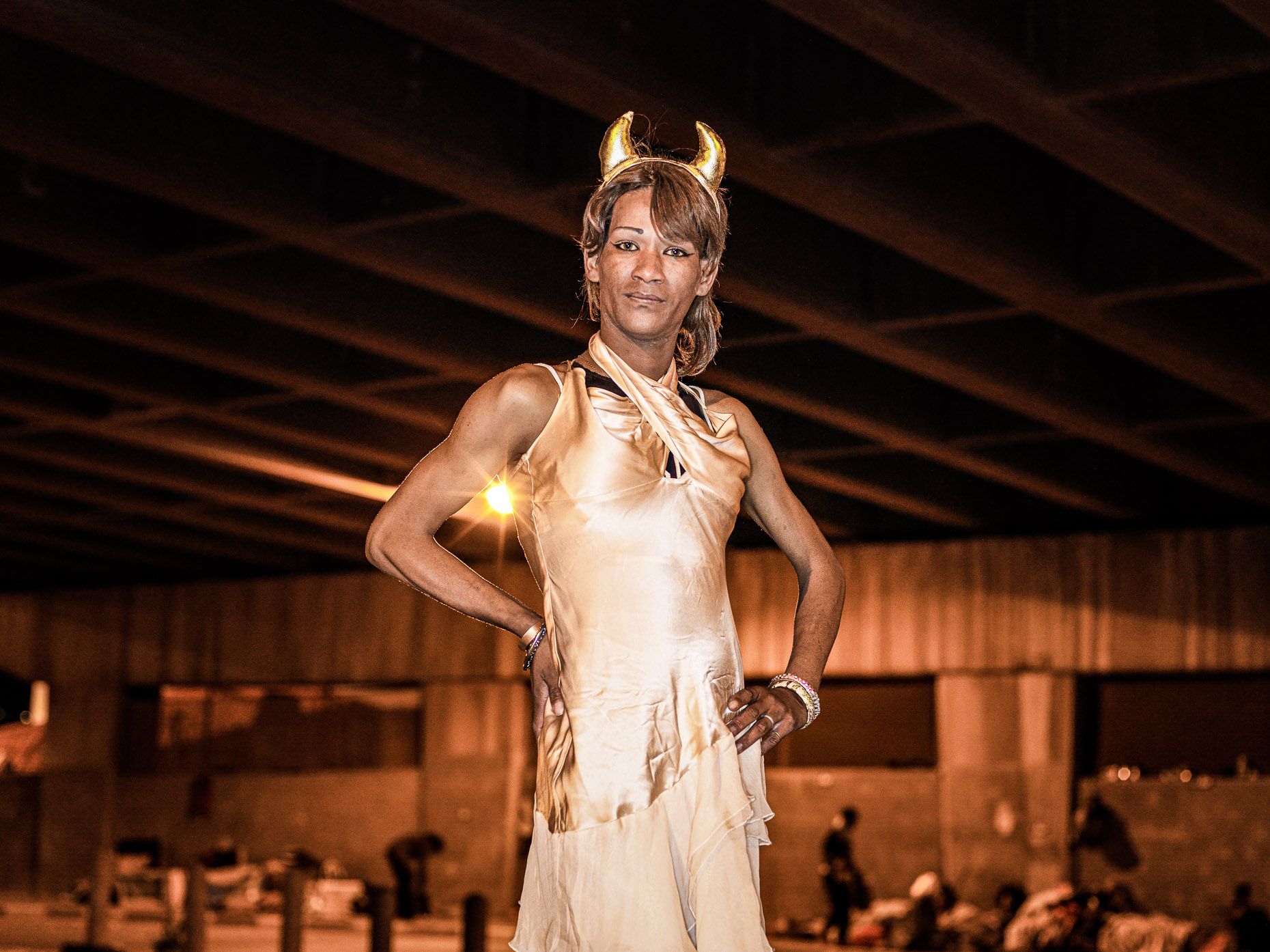 A trans woman, dressed in a gold dress and gold devil horns posing for her portrait. 